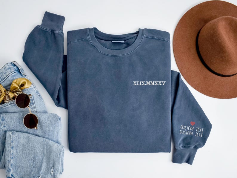 A blue sweatshirt with the text "XXIX MM XXV" printed on it, along with a brown hat and sunglasses, are arranged on a white background.