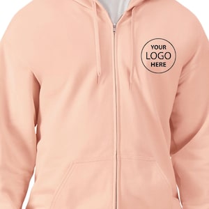 A person wearing a peach-colored hooded sweatshirt with a black logo on the left side of the chest.