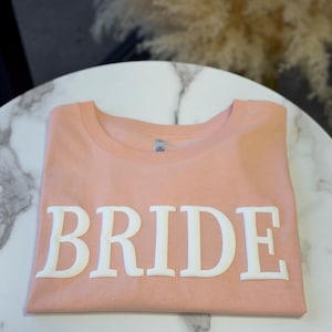 May include: A folded peach-colored t-shirt with the word "BRIDE" in large, white, block letters across the front. The shirt is neatly arranged on a white marble-patterned surface.
