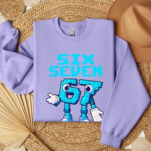 a purple sweatshirt with a graphic design of a character made up of the number 6 and the number 7, along with the text "SIX SEVEN" printed on it. The sweatshirt is laid out on a woven mat, and there is a hat placed next to it.