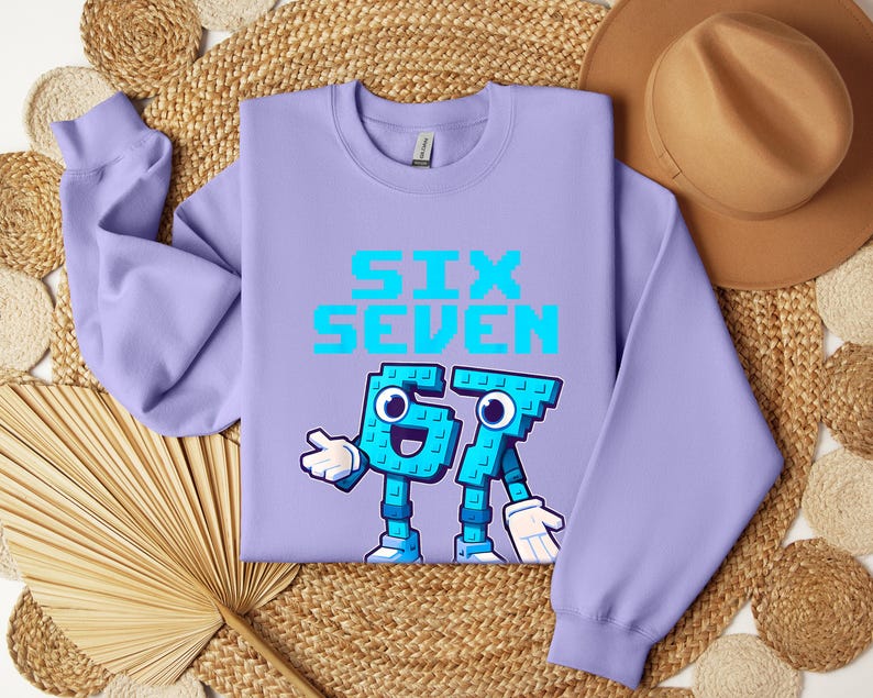 a purple sweatshirt with a graphic design of a character made up of the number 6 and the number 7, along with the text "SIX SEVEN" printed on it. The sweatshirt is laid out on a woven mat, and there is a hat placed next to it.