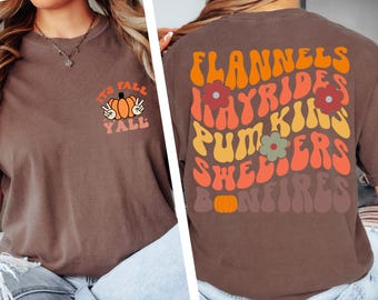May include: Espresso-colored t-shirt with fall-themed graphics. The front features a pumpkin with peace signs and the text "It's Fall Y'all." The back displays the words "Flannels, Hayrides, Pumpkins, Shelters, Bonfires" in a retro font.