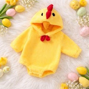 May include: A yellow baby chicken costume with a hood featuring a red comb and beak. The one-piece outfit has long sleeves and a red bow tie. The costume is made of soft, plush material.