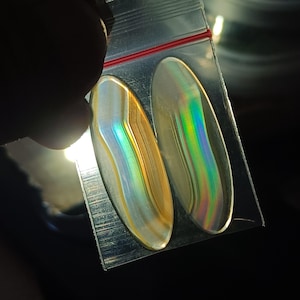 Puede incluir: Dos piedras preciosas ovaladas e iridiscentes se muestran en una bolsa de plástico transparente. Las piedras tienen una apariencia en capas con bandas de tonos dorados, blancos y azules. La bolsa está sellada con una cremallera roja.