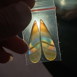 Puede incluir: Dos piedras preciosas en forma de lágrima con bandas iridiscentes de color, sujetas en una bolsa de plástico transparente. Las piedras tienen un aspecto pulido y translúcido, con tonos amarillos, naranjas y toques de azul y verde. La luz brilla a través de las piedras.