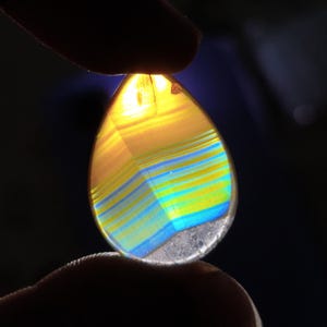 Può includere: Una gemma a forma di goccia con strati di tonalità gialle, blu e verdi. La pietra è tenuta tra due dita, con la luce che la attraversa, evidenziando le bande di colore. La parte inferiore della pietra è bianca traslucida.