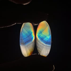 Puede incluir: Dos cabujones de ágata iridiscente de forma ovalada con un patrón de color azul, amarillo y blanco.