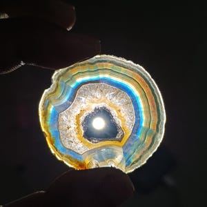 May include: A translucent, circular agate slice with concentric bands of color, including blue, yellow, and orange. The center features a dark, irregular opening, with light shining through the stone. The agate is held between two fingers against a dark background.