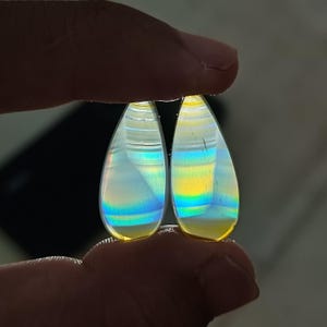 Puede incluir: Dos piedras preciosas en forma de lágrima con colores arcoíris iridiscentes. Las piedras se sostienen entre los dedos, mostrando sus superficies pulidas y el juego de luces interno. Las gemas tienen un brillo nacarado, con tonos de azul, amarillo y verde.