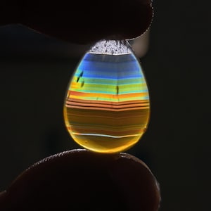 May include: A teardrop-shaped gemstone featuring horizontal bands of color including blue, green, orange, and yellow. The translucent stone is backlit, highlighting the layered pattern and smooth, polished surface of the faceted, clear, and amber-toned material.