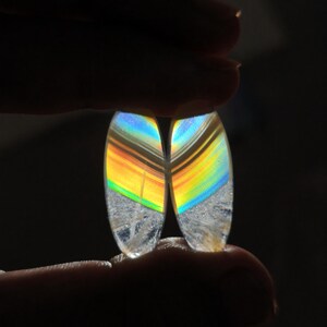Puede incluir: Dos piedras preciosas ovaladas con iridiscencia de arcoíris. Las piedras se sostienen sobre un fondo oscuro, mostrando el juego de la luz en sus superficies. La parte inferior de cada piedra es un material cristalino transparente.