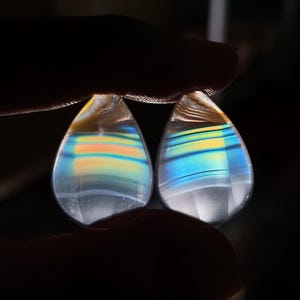 May include: Two teardrop-shaped, iridescent gemstones with bands of yellow, orange, and blue. The stones are held between fingers against a dark background, showcasing their translucent quality and vibrant colors. These could be used as earrings or pendants.