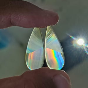 Puede incluir: Dos gemas claras en forma de lágrima se sostienen entre los dedos. Las gemas reflejan un arcoíris de colores, con una sección blanca semitransparente en la parte superior. La luz se refracta a través de las gemas, creando un efecto de prisma.