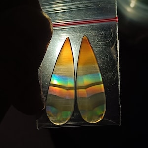Puede incluir: Dos piedras preciosas en forma de lágrima con colores arcoíris iridiscentes. Las piedras tienen un degradado de tonos naranja, amarillo y transparente. Las piedras preciosas se muestran en una bolsa de plástico transparente con un sello rojo.