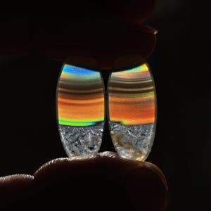 Puede incluir: Dos piedras preciosas ovaladas con un diseño colorido y en capas. Las piedras presentan bandas de tonos naranja, amarillo y azul, que recuerdan a una puesta de sol. Las secciones inferiores tienen una apariencia cristalina y texturizada.