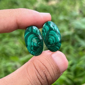 May include: Two oval malachite gemstones. The stones are a vibrant green with intricate, swirling patterns of darker green. The polished stones are held against a blurred green background.