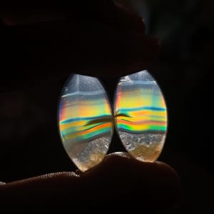May include: Two oval gemstones with a rainbow iridescence. The stones show bands of yellow, orange, green, and blue, with a clear, crystalline appearance. The gemstones are held between fingers against a dark background.