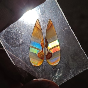 Puede incluir: Dos piedras preciosas en forma de lágrima con una mezcla de colores, incluyendo marrón, azul y naranja. Las piedras tienen una apariencia en capas y están sobre una superficie reflectante. Las piedras preciosas son probablemente para la fabricación de joyas.