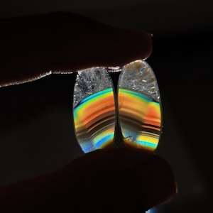 Puede incluir: Dos piedras preciosas ovaladas se sostienen entre los dedos sobre un fondo oscuro. Las piedras muestran un arcoíris de colores, incluyendo naranja, amarillo, verde y azul, con bandas oscuras. La parte superior de cada piedra es transparente.
