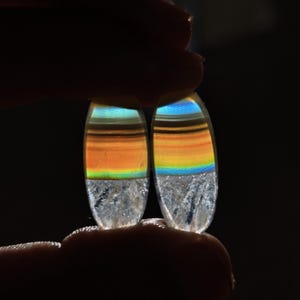 Puede incluir: Dos piedras preciosas ovaladas con un arcoíris de colores, incluyendo naranja, amarillo, verde y azul, se sostienen entre los dedos. La parte inferior de cada piedra tiene una apariencia clara y cristalina.