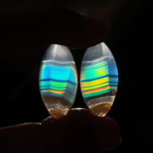 May include: Two oval-shaped gemstones with iridescent bands of blue, green, yellow, and orange. The stones have a translucent appearance with a light brown base and are held against a black background.