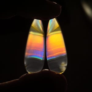 May include: Two teardrop-shaped iridescent agate cabochons with a rainbow of colors, including yellow, orange, blue, and green.