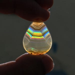 May include: A teardrop-shaped translucent gemstone featuring horizontal bands of iridescent rainbow colors. The stone is backlit, highlighting the internal layers and the spectrum of light passing through the clear, amber-toned material.