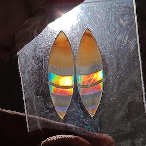 Puede incluir: Dos gemas alargadas en forma de lágrima. La parte superior es de color marrón claro, que pasa a una banda gris con una vibrante iridiscencia de arcoíris. La parte inferior es clara y translúcida. Las gemas se muestran sobre una superficie reflectante.