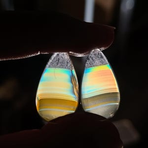 May include: Two teardrop-shaped gemstones with iridescent bands of colour, including yellow, orange, and blue. The stones are held between fingers against a dark background, highlighting their polished surfaces and natural patterns. The top of each stone has a crystalline texture.