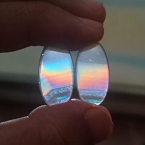 Puede incluir: Dos gemas ovaladas e iridiscentes se sostienen entre los dedos. Muestran un arcoíris de colores, incluyendo azul, naranja y amarillo, con una base texturizada y cristalina. Las gemas son probablemente para la fabricación de joyas.