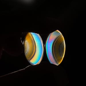 May include: Two oval-shaped gemstones with iridescent rainbow colors. The stones have a polished surface and display bands of yellow, blue, and purple hues. The background is black, highlighting the gemstones' vibrant colors.