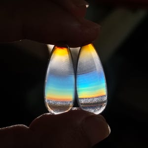 Puede incluir: Dos gemas en forma de lágrima con colores del arcoíris, desde el amarillo hasta el azul. Se sostienen sobre un fondo oscuro, resaltando sus cualidades iridiscentes. La base de cada piedra tiene una apariencia texturizada y gris.