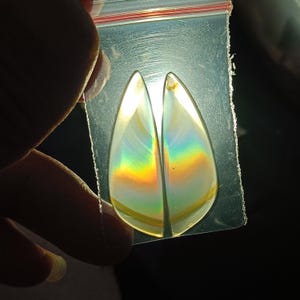 Puede incluir: Dos piedras preciosas iridiscentes en forma de lágrima se sostienen en una bolsa de plástico transparente. Las piedras muestran un arcoíris de colores, incluyendo amarillo, naranja, verde y azul. Las piedras son lisas y pulidas.