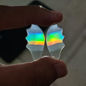 Puede incluir: Dos objetos translúcidos iridiscentes en forma de alas de mariposa. Las alas muestran un espectro de colores, incluyendo verde, amarillo y naranja, que reflejan la luz. Las alas se sostienen entre los dedos, con un fondo oscuro.