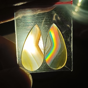 Puede incluir: Dos piedras translúcidas en forma de lágrima se muestran en una bolsa de plástico transparente. Una piedra tiene un degradado de amarillo y marrón, mientras que la otra refracta la luz en un arcoíris de colores. Las piedras se sostienen sobre un fondo oscuro.