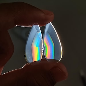 Puede incluir: Dos gemas claras en forma de lágrima se sostienen a la luz, mostrando un vibrante espectro de arcoíris en su interior. Las gemas están facetadas, capturando y refractando la luz para crear el efecto colorido. El fondo es gris oscuro.