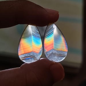 May include: Two teardrop-shaped, iridescent gemstones. The stones display a spectrum of colors, including orange, yellow, blue, and green, with a clear, polished surface. The gemstones are held up by a person's fingers.