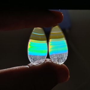Puede incluir: Dos gemas iridiscentes en forma de lágrima. Las piedras tienen capas de amarillo, azul y verde, con una base transparente y texturizada. Las gemas se sostienen entre los dedos, mostrando sus colores vibrantes y patrones únicos.