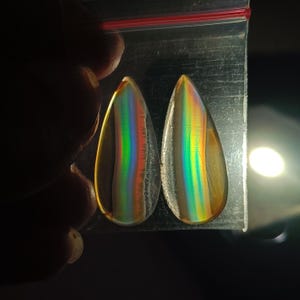 Puede incluir: Dos piedras preciosas iridiscentes en forma de lágrima se muestran en una bolsa de plástico transparente. Las piedras tienen un efecto arcoíris con bandas de rojo, naranja, amarillo, verde y azul. Las piedras se sostienen en una mano.