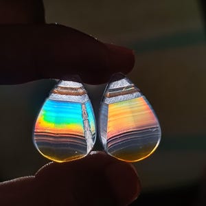 May include: Two teardrop-shaped, iridescent gemstones. The stones display a spectrum of colors, including blue, green, orange, and yellow, with horizontal banding. The top of each stone has a silver-colored band. The gemstones are held between fingers.