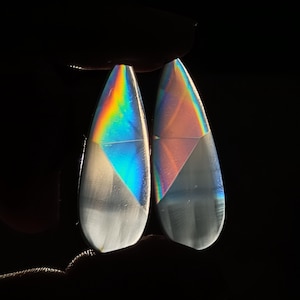Puede incluir: Dos piedras iridiscentes en forma de lágrima con un patrón blanco y gris. Las piedras tienen un brillo arcoíris.