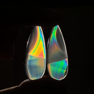 Puede incluir: Dos cabujones de ágata iridiscente, cada uno con forma de lágrima y un arcoíris de colores, incluyendo azul, verde, amarillo y naranja.