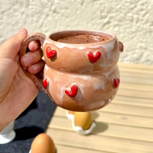 Könnte beinhalten: Ein runder, bauchiger Keramikbecher mit einer glänzenden hellbraunen Glasur und einem weißen Tropfmuster am Rand. Der Becher ist mit mehreren kleinen, erhabenen, leuchtend roten herzförmigen Verzierungen auf der Außenseite und einem Henkel versehen.