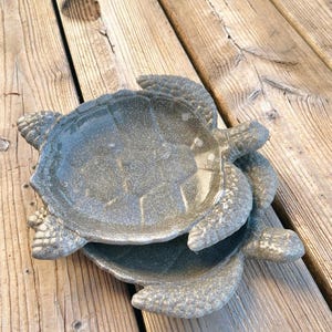 Puede incluir: Dos platos decorativos grises con forma de tortuga apilados uno encima del otro. Los platos tienen una superficie texturizada y están colocados sobre una terraza de madera. Los platos de tortuga son un artículo de decoración único.