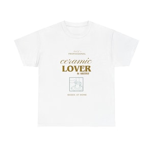 Könnte beinhalten: Ein weißes Kurzarm-T-Shirt mit goldenem und dunkelblauem Text, der "kind of a PROFESSIONAL ceramic LOVER & ARTIST" lautet, sowie einer kleinen Strichzeichnung einer Person, die einen Tontopf formt, und dem Text "BASED: AT HOME" darunter.