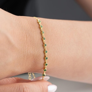 May include: Gold-toned bracelet featuring a delicate chain adorned with small, round, green gemstones. The bracelet is displayed on a wrist, showcasing its elegant design and subtle sparkle. A piece of jewelry suitable for everyday wear or special occasions.