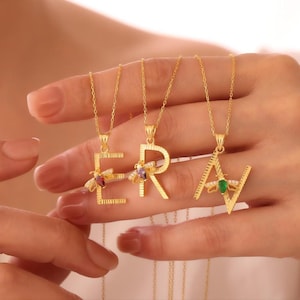 Puede incluir: Collares iniciales dorados que deletrean la palabra "ERA". Cada colgante de letra presenta un pequeño diseño de insecto colorido. Los collares se muestran en la mano de una persona.