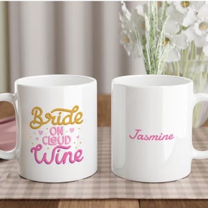 Könnte beinhalten: Zwei weiße Keramikbecher. Ein Becher hat den Text "Bride on Cloud Wine" in Gold und Rosa. Der andere Becher hat den Namen "Jasmine" in Rosa. Beide Becher haben einen gebogenen Griff.