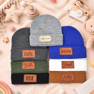 May include: A collection of seven knit beanies in various colors including gray, dark gray, olive green, black, blue, white, and tan. Each beanie features a rectangular leather-like patch with a name engraved in black text: Anna, Katie, Bella, Rosie, Becky, Sarah, and Kelly.