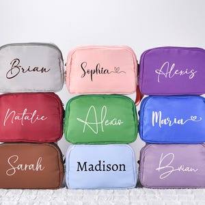 Könnte beinhalten: Neun personalisierte Kosmetiktaschen in verschiedenen Farben, darunter Grau, Rosa, Lila, Rot, Grün, Blau und Braun. Jede Tasche trägt einen Namen in eleganter Schrift. Die Taschen sind für die Aufbewahrung von Make-up und Toilettenartikeln konzipiert.